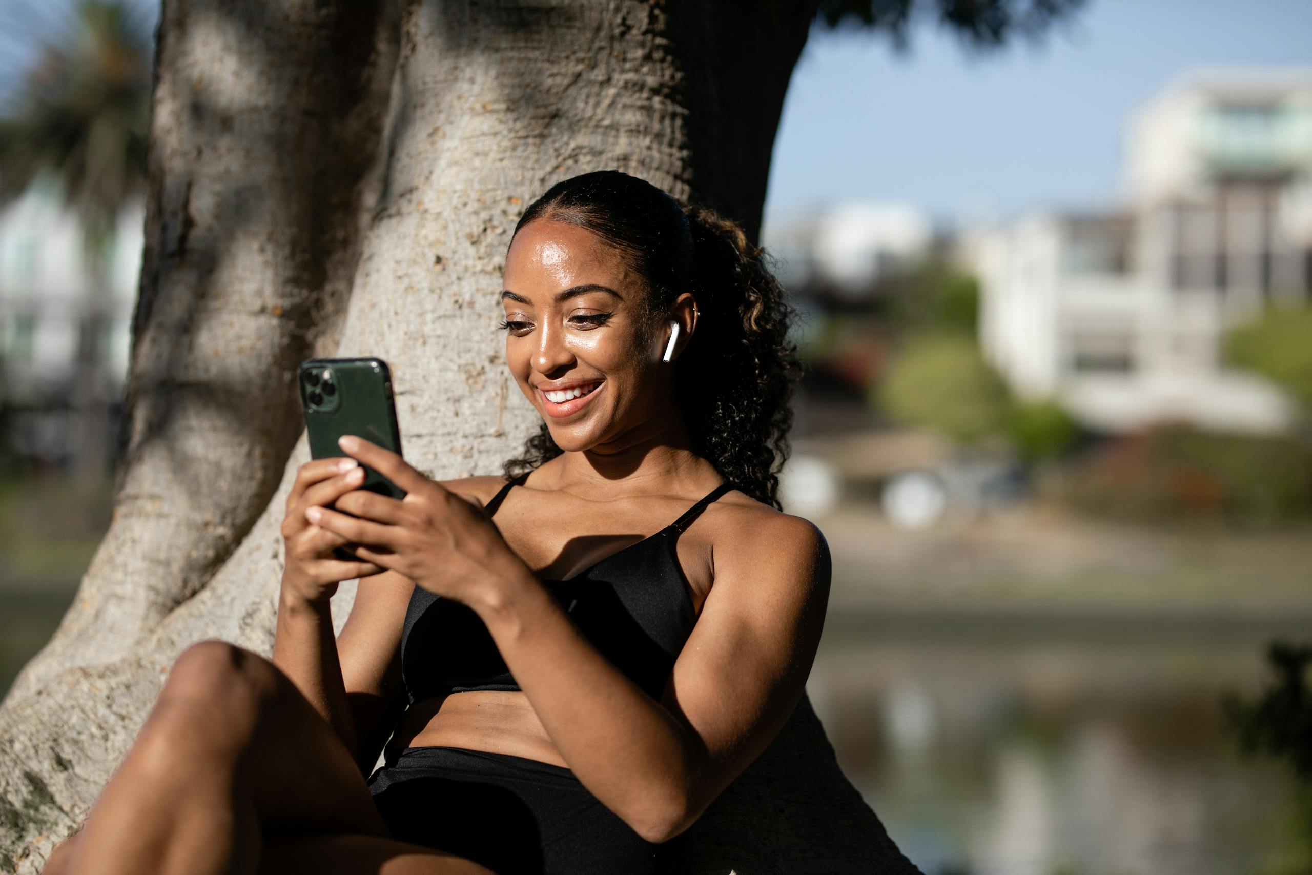 A happy woman sitting under a tree, texting on her smartphone while enjoying the sunny outdoors.