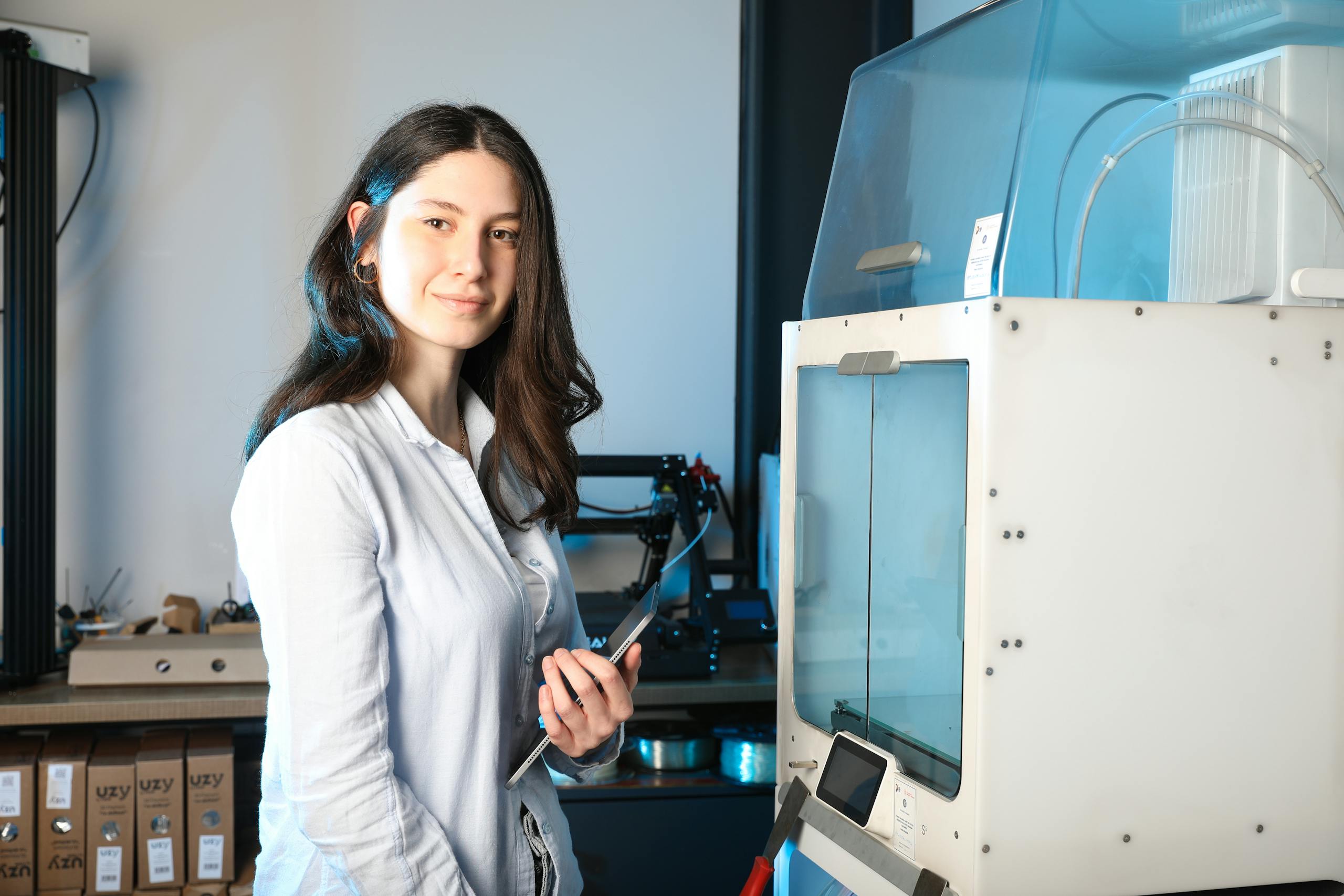 Confident young female engineer holding a tablet beside a 3D printer in a modern indoor setting.