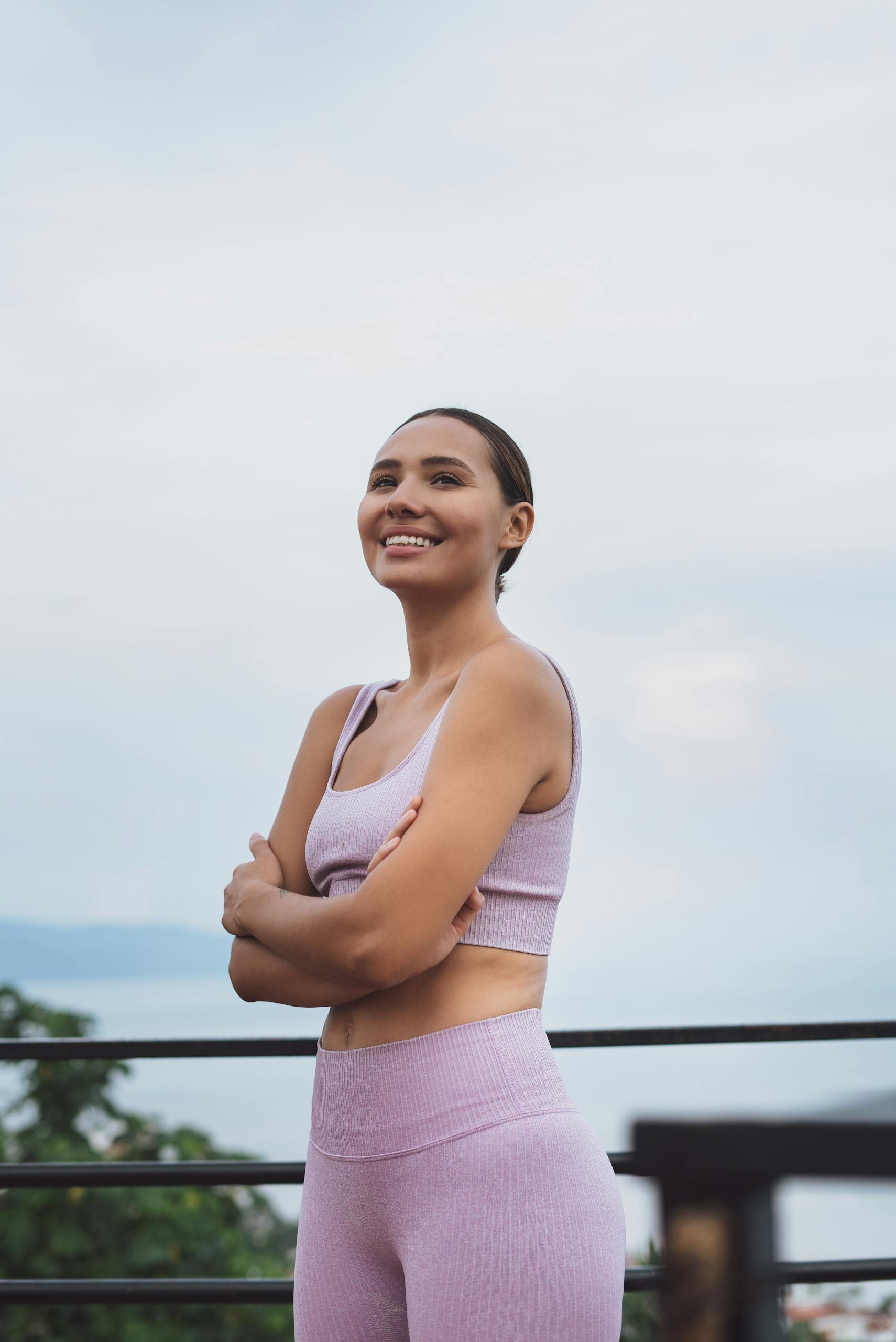 Smiling woman with arms crossed, wearing a sports bra and yoga pants, standing outdoors.
